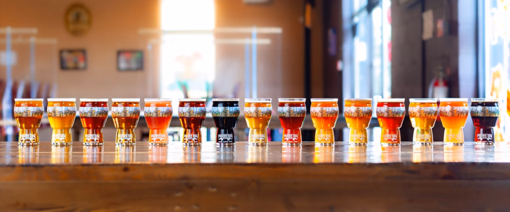 15 Prairie Dog Brewing craft beers arranged in a long row with 16-oz beer glasses.