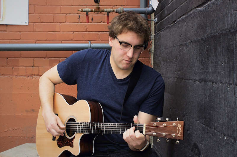 Calgary musician Grant Kanigan plays his guitar against a corner of rend and black brick walls.