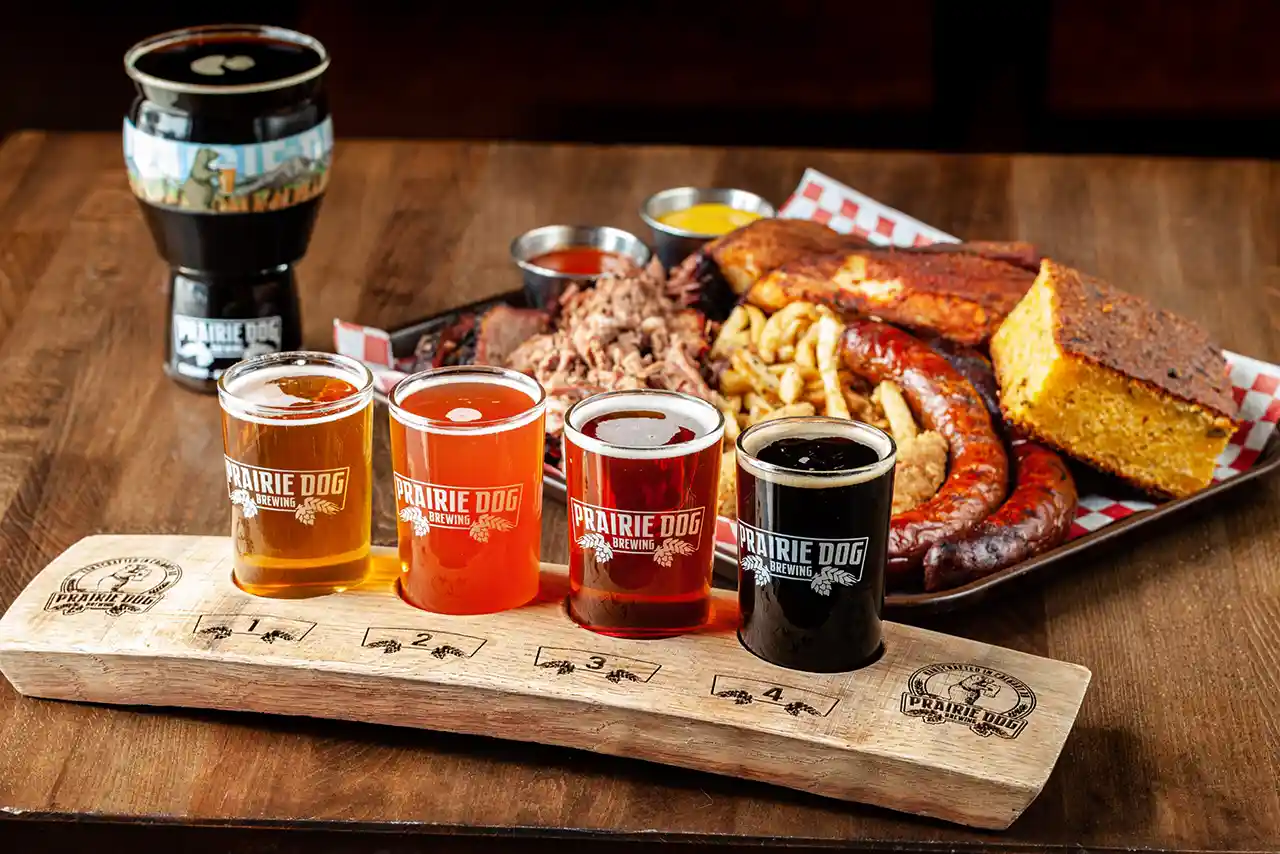 A flight of 4 Prairie Dog Brewing craft beers in branded glassware on a wooden paddle, with a 16oz pint of stout in the background, and a platter of delicious BBQ including sausages, chicken tenders, and beef brisket.